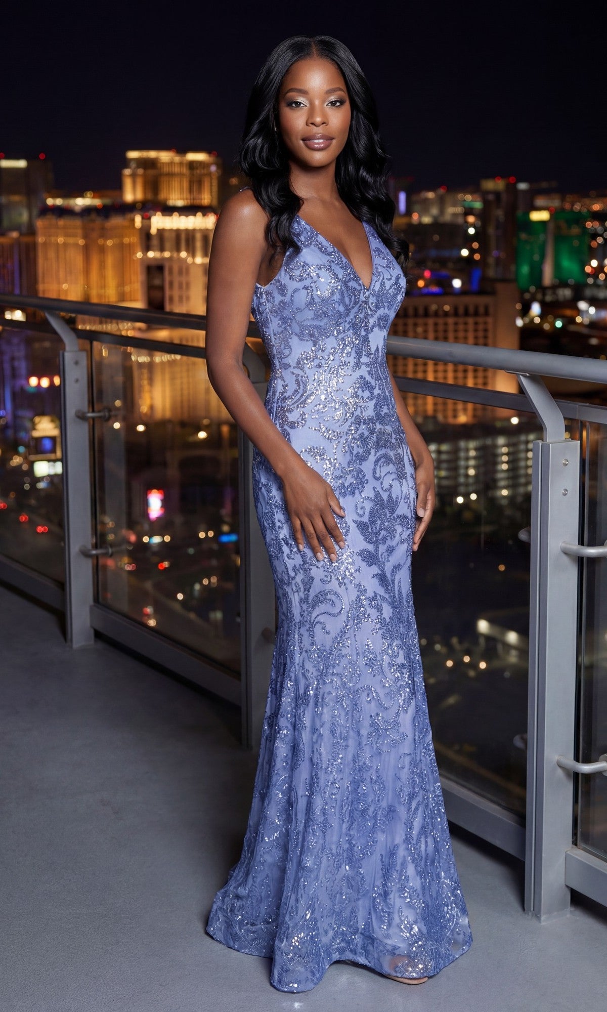 Woman in a sleeveless, floor-length Blondie Nites 4501BN long sequin-print prom dress in periwinkle stands on a balcony at night with city lights behind her.